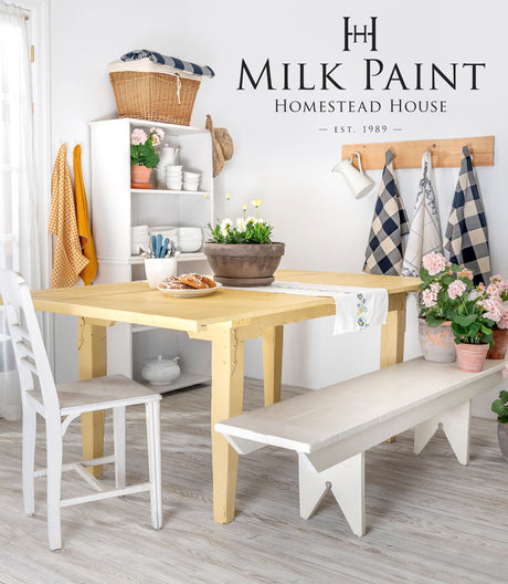Dining table in Homestead House’s Swedish Yellow Milk Paint with white bench and plants nearby.