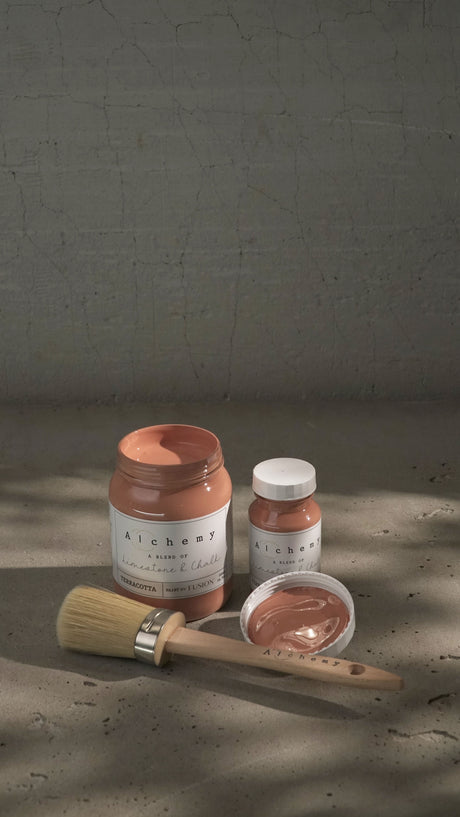 Three Terracotta Alchemy Paint by Fusion cans with a paintbrush on concrete in soft light.
