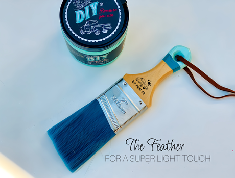 The Feather DIY Paintbrush by DIY Paint Co., blue tip facing up, on a white surface near paint.