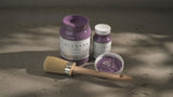 Two jars of Thistle Alchemy Paint by Fusion, one open with a brush resting nearby on a concrete surface.