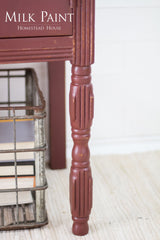 Table leg painted in Homestead House Trading Post Red Milk Paint on a textured rug near books.