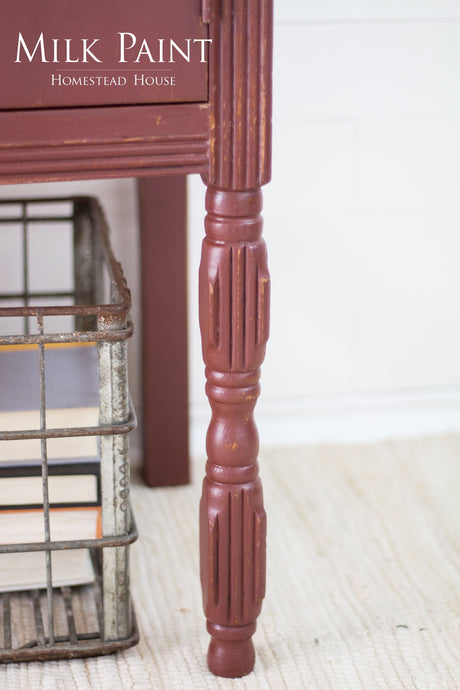 Table leg painted in Homestead House Trading Post Red Milk Paint on a textured rug near books.