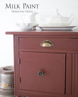 Cabinet in Homestead House Trading Post Red Milk Paint with brass handles, topped with white dishes and a barrel nearby.