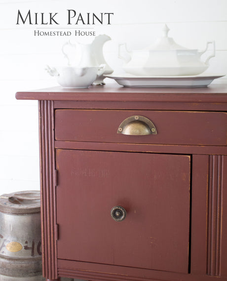 Cabinet in Homestead House Trading Post Red Milk Paint with brass handles, topped with white dishes and a barrel nearby.