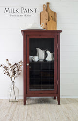 Cabinet painted Trading Post Red Milk Paint by Homestead House, with dishes inside; boards and vase nearby.