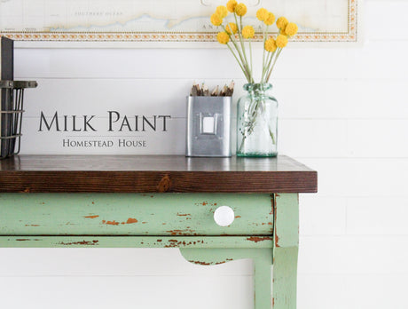 Distressed table in Upper Canada Green Milk Paint with brown top, yellow flower vase, and pencil box.