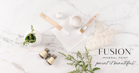 Fusion Mineral Paint Victorian Lace (multi-tone white) with paintbrushes, small plant, and binoculars on marble.