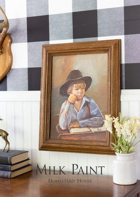 Framed boy-in-black-hat artwork on desk with books and flowers; Homestead House Voyageur Milk Paint info shown below.