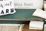 Distressed bench painted in Waterloo Green Milk Paint by Homestead House, styled with books, pillow, and branding.