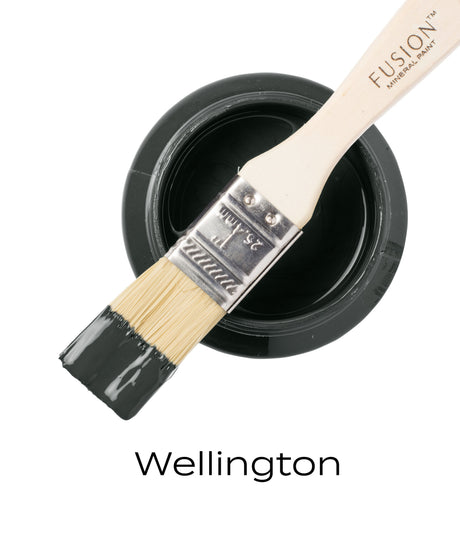 Paintbrush with green-grey on open Wellington Fusion Mineral Paint jar; brand name visible in black text below.