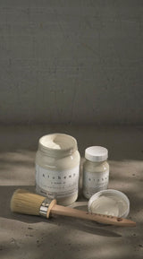 Two jars of White Oak Alchemy Paint by Fusion Mineral Paint with a brush on a softly lit surface.