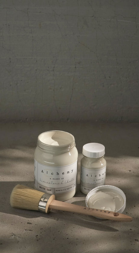 Two jars of White Oak Alchemy Paint by Fusion Mineral Paint with a brush on a softly lit surface.