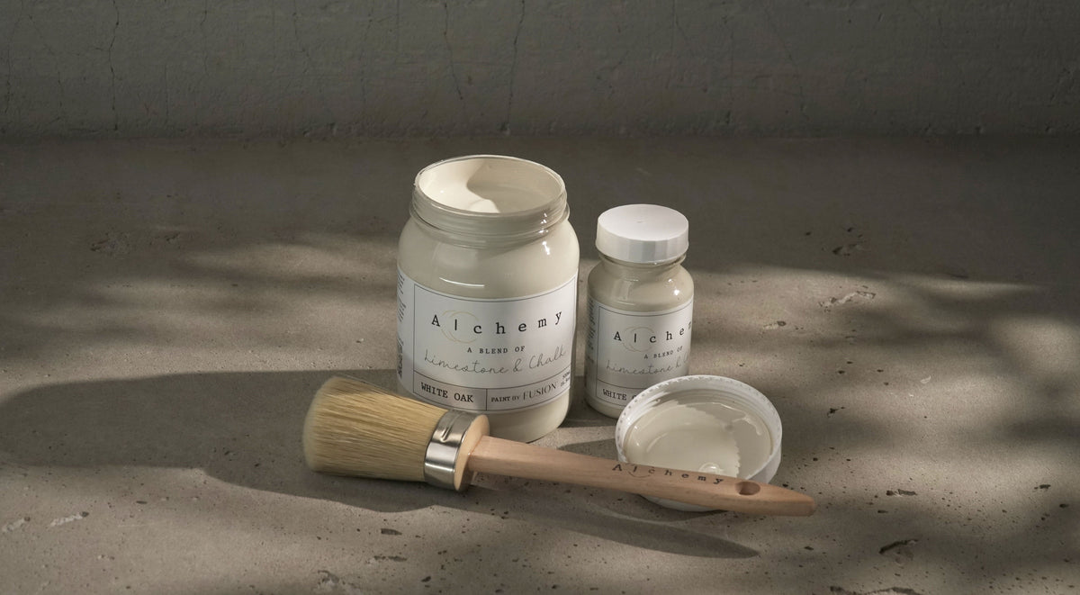 Three White Oak Alchemy Paint jars by Fusion Mineral Paint on gray surface, one jar open with a brush.