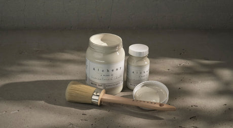 Three White Oak Alchemy Paint jars by Fusion Mineral Paint on gray surface, one jar open with a brush.