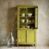 Green wooden cabinet in Willow Alchemy Paint by Fusion Mineral Paint holding white dishes, vase with branches beside it.