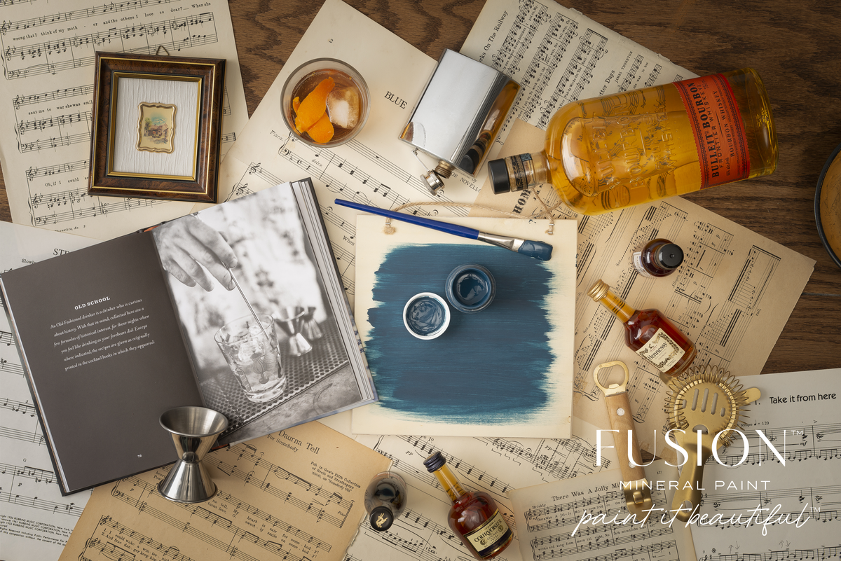 Willowbank Fusion Mineral Paint jar on a wooden table with brushes, books, whiskey, and sheet music nearby.