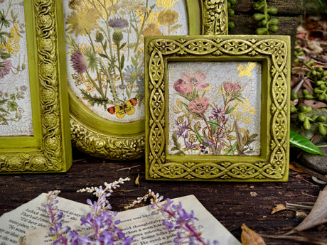 Two gold frames with botanical art on an IOD Wood Gallery Blank by I.O.D., shown beside a book and purple flowers.