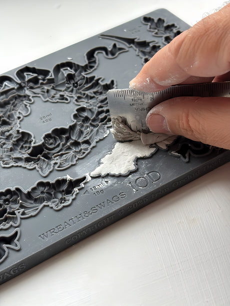 Hand pressing clay into IOD Wreath & Swags Mould with a ruler on a white surface.
