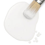 Paintbrush in spilled Fusion Mineral Paint Hotel Robe Milk Paint, bristles coated white with small droplets nearby.